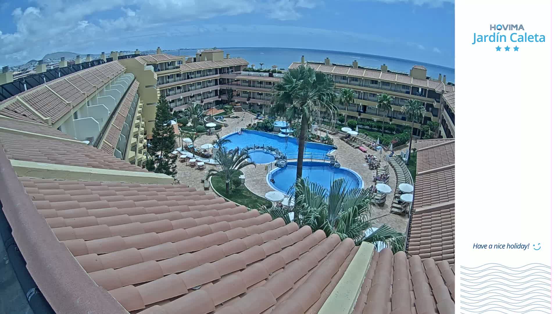 An overhead view on a sunny day shows a resort complex with multiple pools surrounded by buildings and palm trees, overlooking a calm ocean.