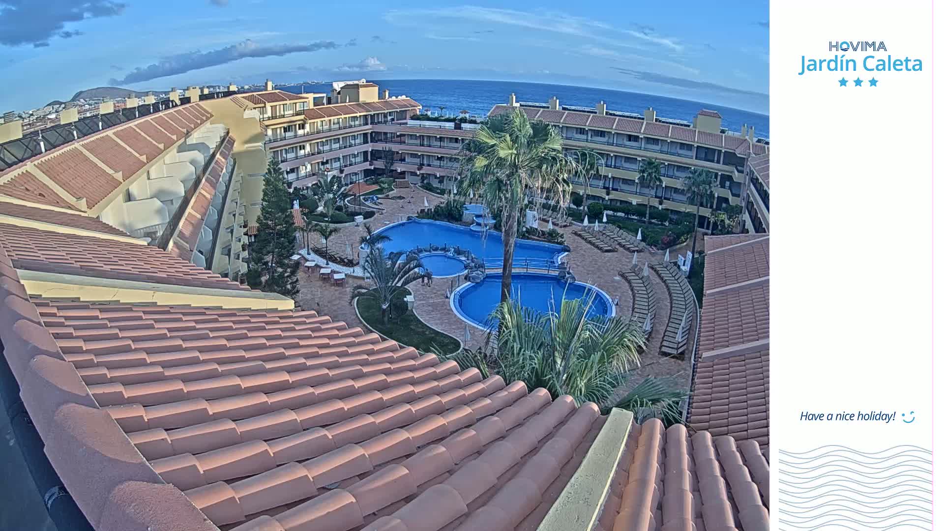 A view from a rooftop shows a resort courtyard with two swimming pools, palm trees, and surrounding buildings under a partly cloudy sky.