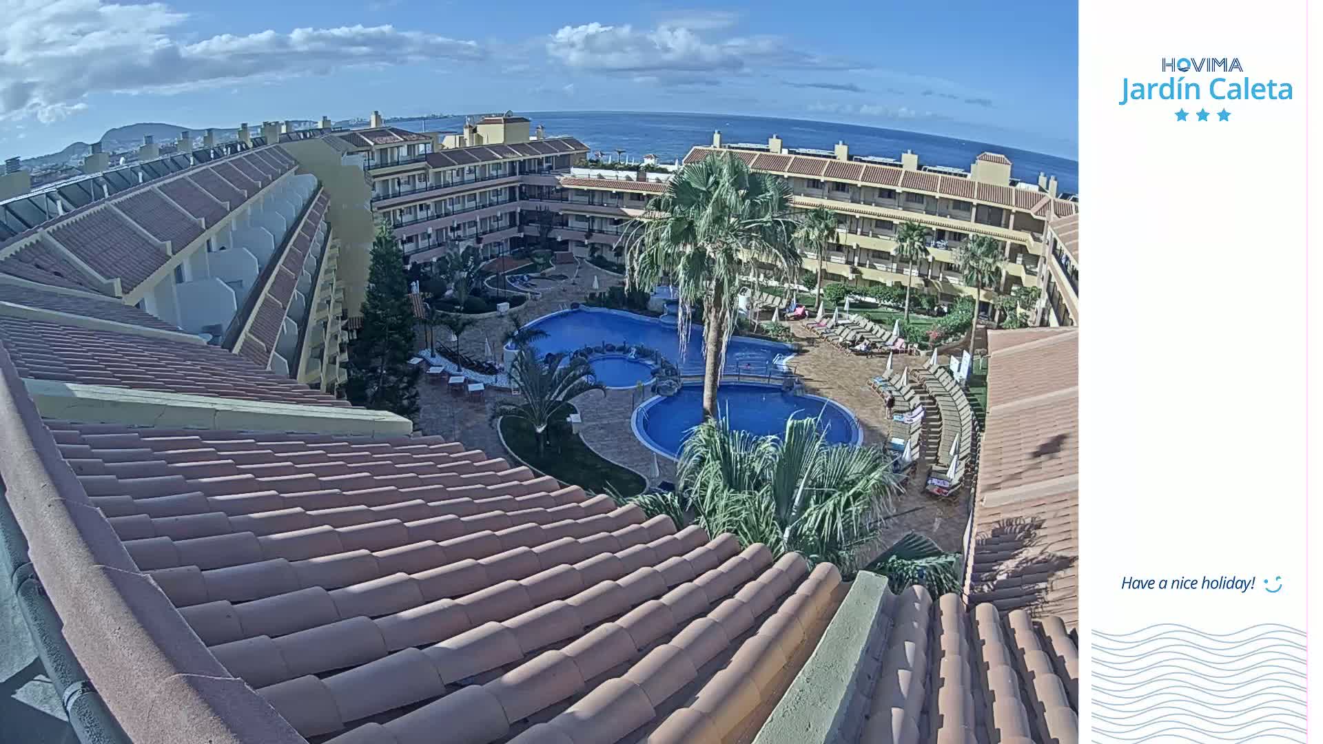 A tiled roof overlooks a resort courtyard with multiple pools, palm trees, and sun loungers, all under a partly cloudy sky.
