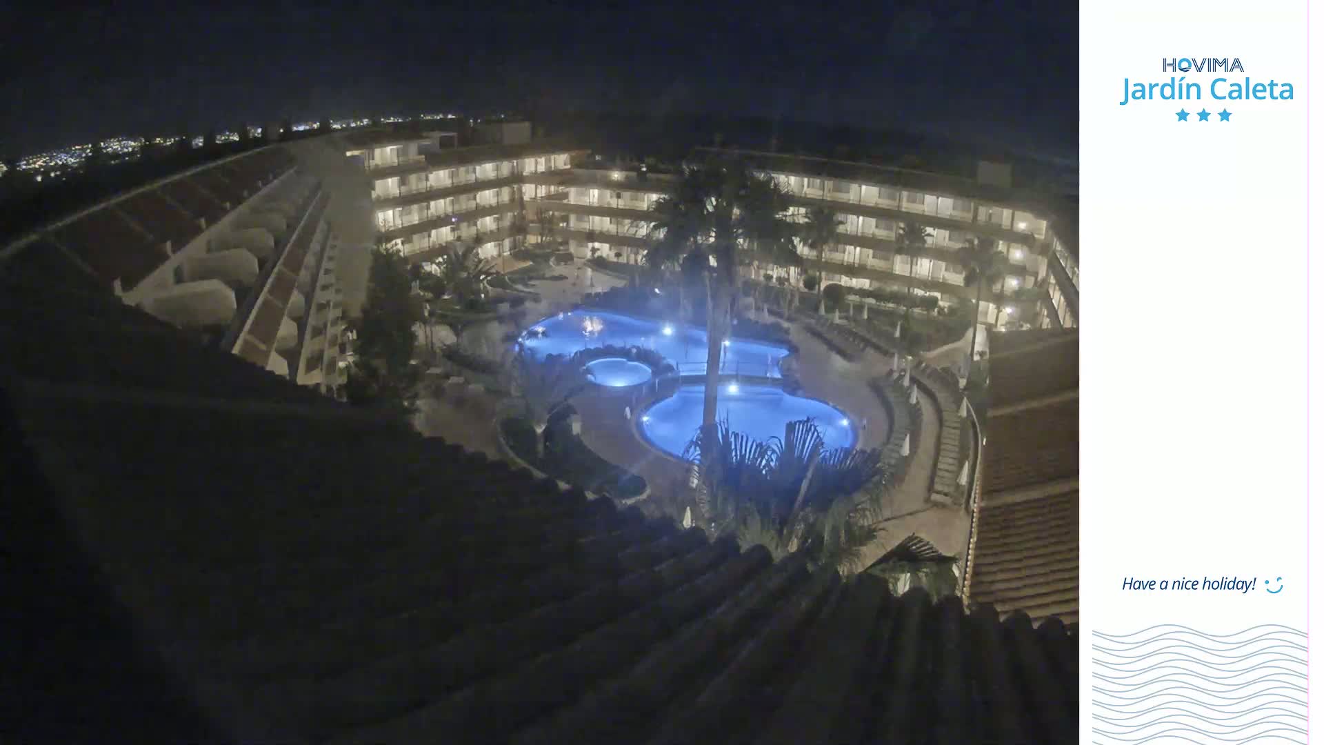 A nighttime aerial view shows a multi-story building complex surrounding a lit swimming pool area with palm trees.