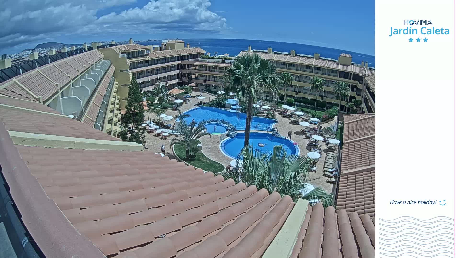 A partly cloudy day overlooks a resort with terracotta roofs, multiple swimming pools, and palm trees surrounding a courtyard.
