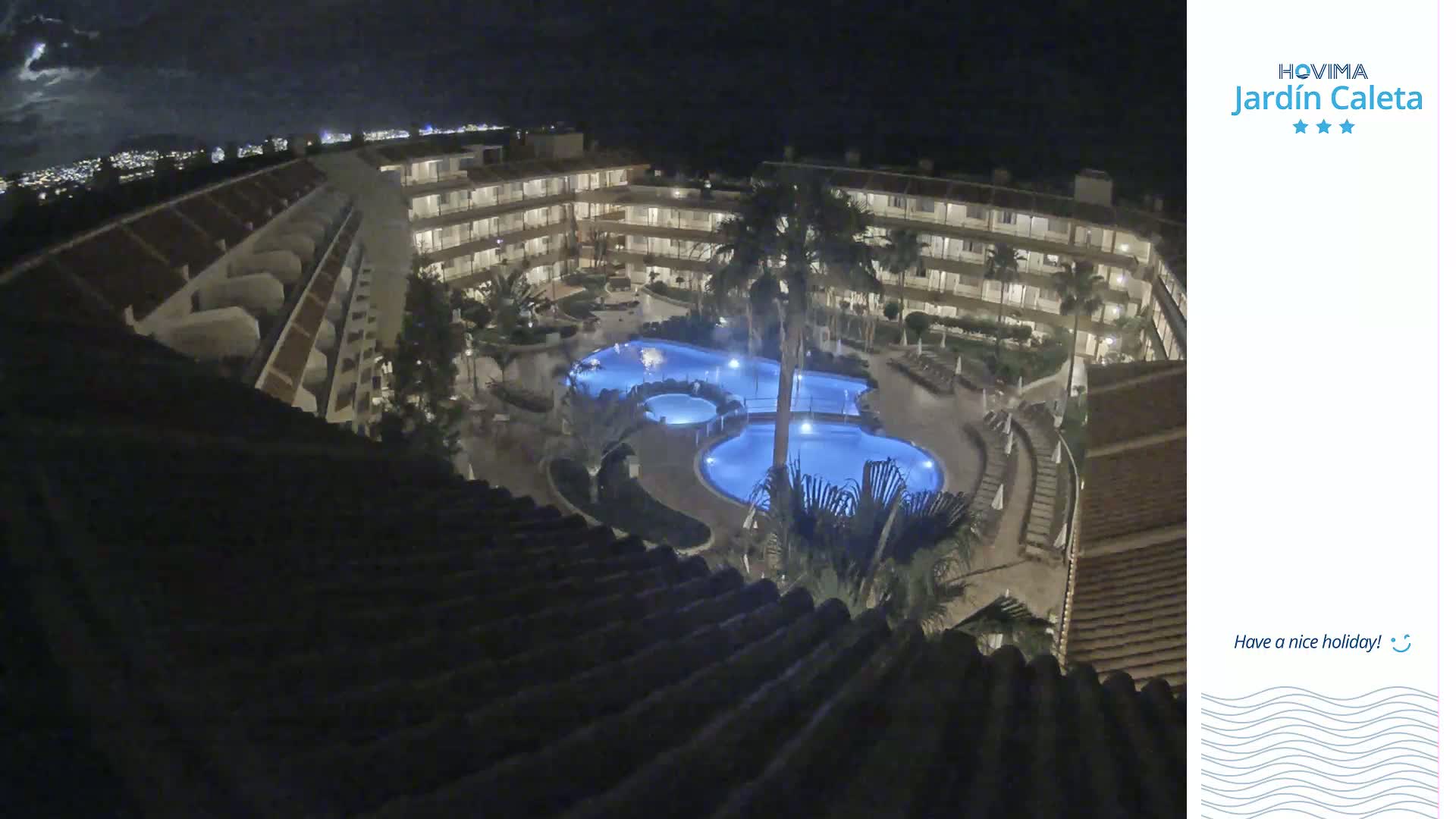 A nighttime aerial view shows a multi-story building complex surrounding two illuminated pools, with palm trees and landscaping visible under a partly cloudy sky.