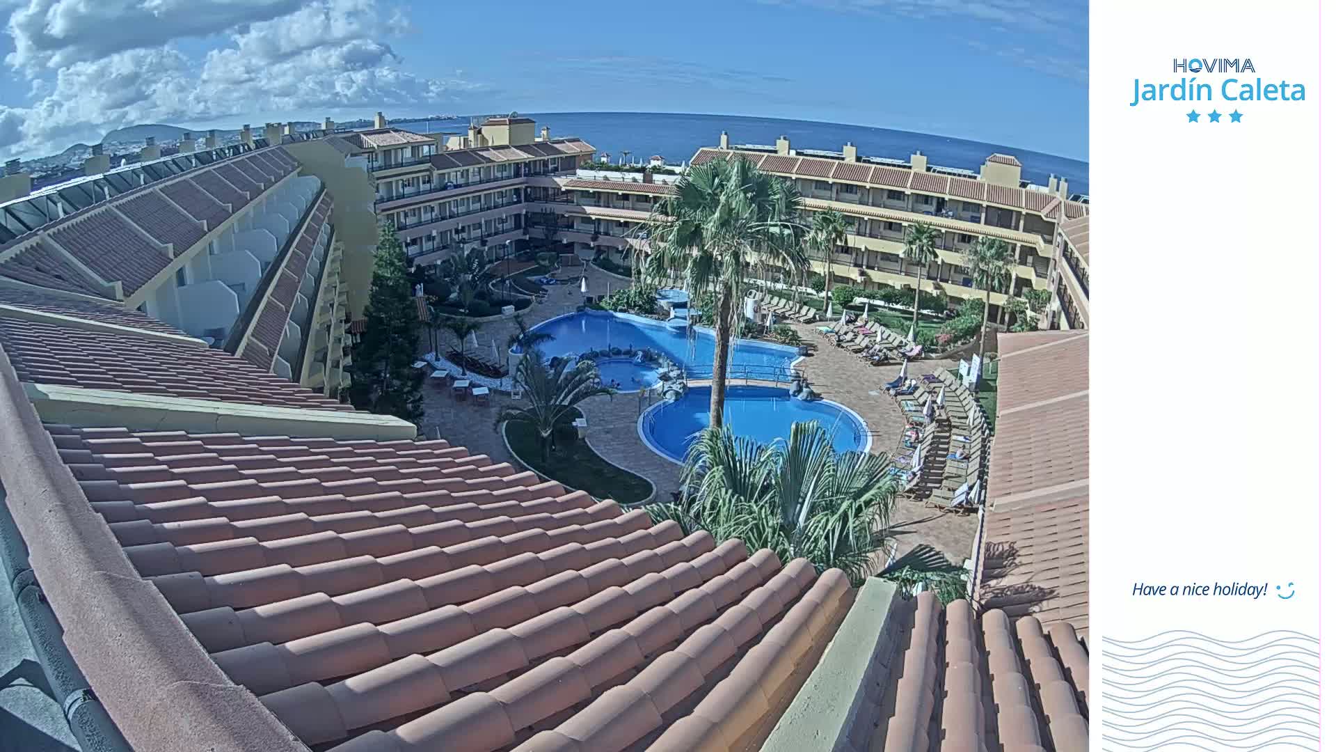 An overhead view of a resort complex with multiple pools and surrounding buildings, on a sunny day with a clear view of the ocean in the distance.