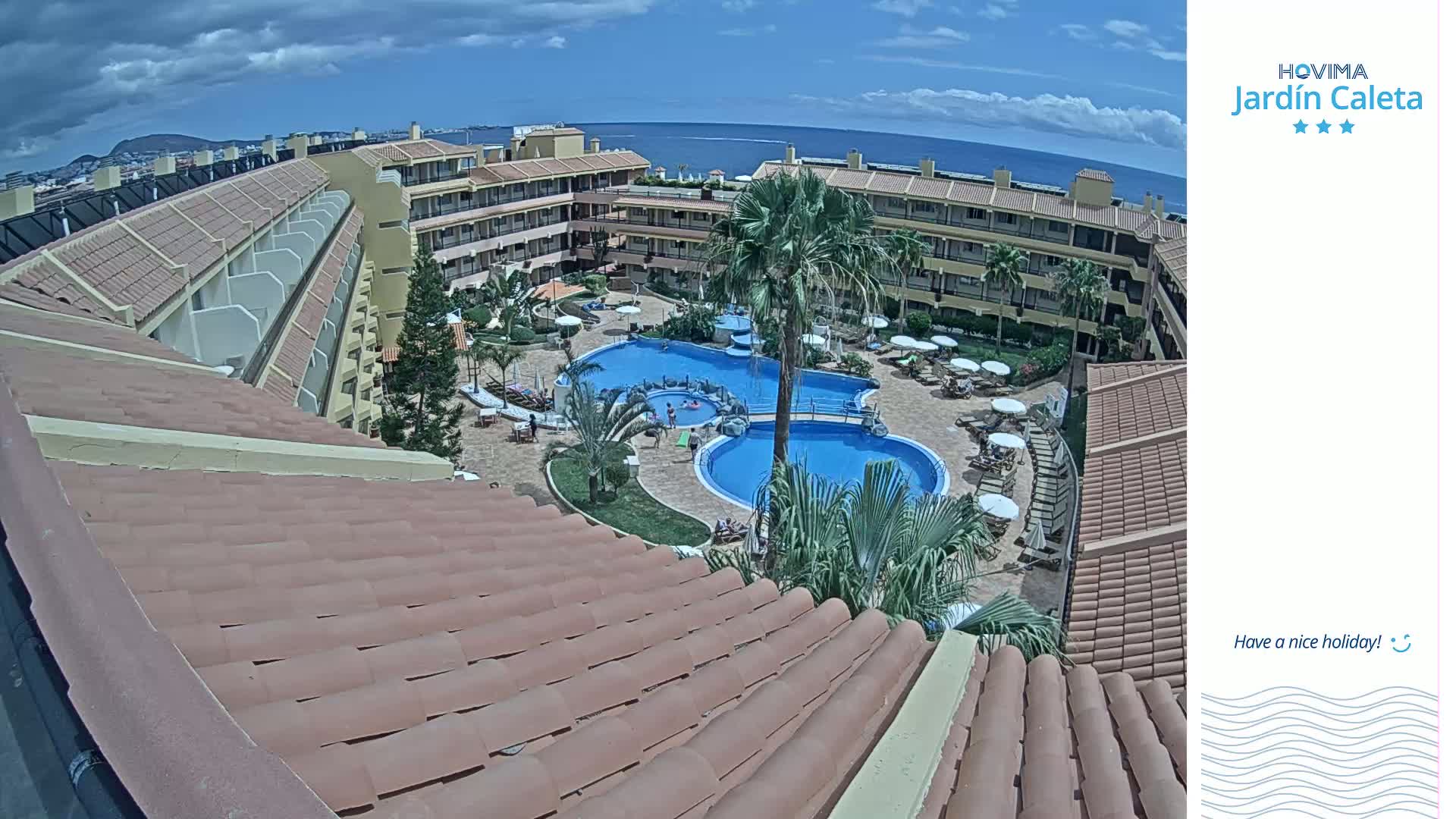 An aerial view on a sunny day shows a resort complex with two swimming pools surrounded by palm trees and lounge chairs, partially visible from a tiled roof.