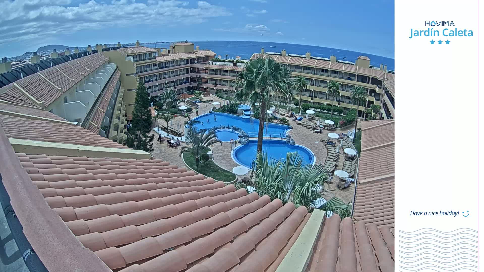 An overhead view of a resort with multiple pools, surrounded by buildings under a partly cloudy sky and overlooking a calm ocean.