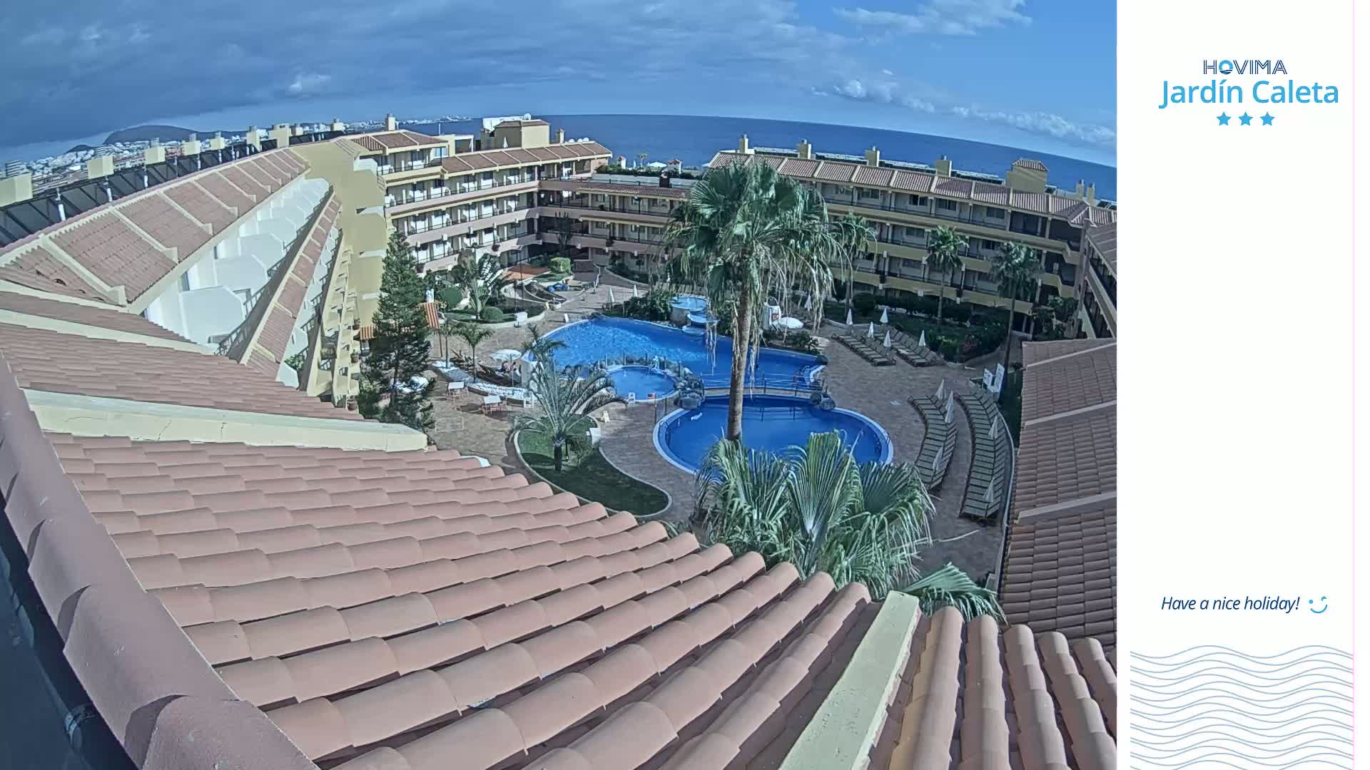 An aerial view of a resort complex with multiple pools, palm trees, and a tiled roof under a partly cloudy sky.