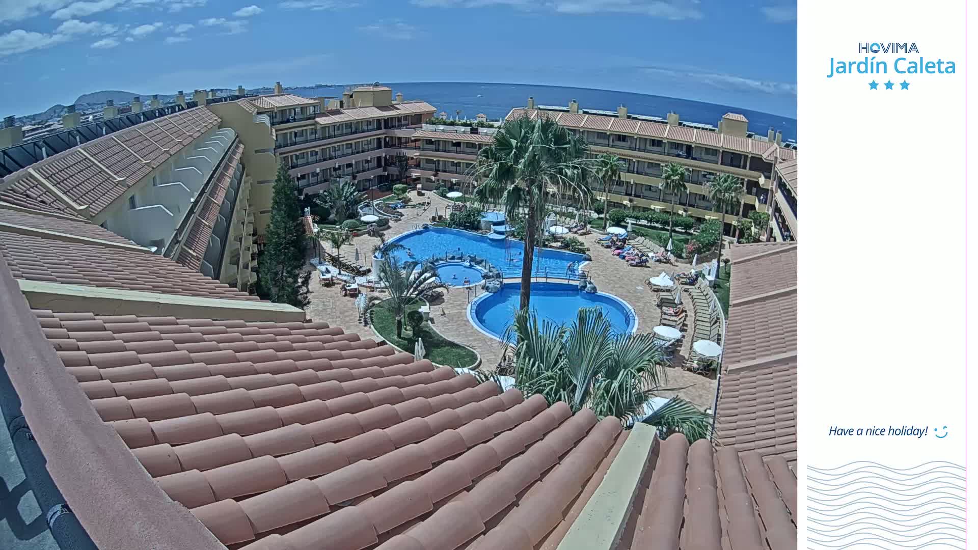 A rooftop view of a resort complex on a sunny day shows a courtyard with two swimming pools surrounded by buildings and lounge chairs.