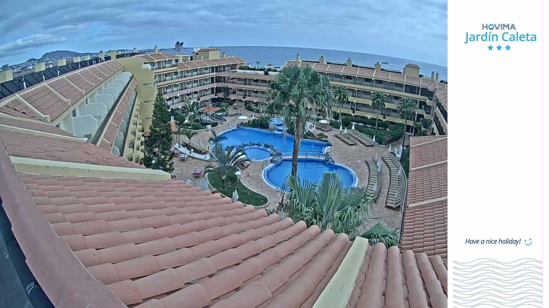 An aerial view shows a resort with multiple buildings surrounding a courtyard containing two swimming pools under an overcast sky.