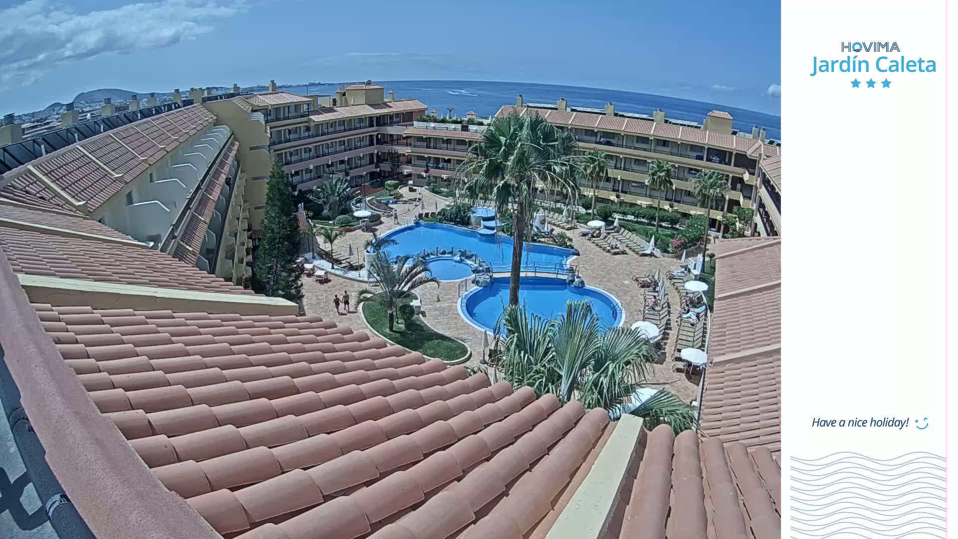 A sunny day overlooks a resort's courtyard with two swimming pools, surrounded by buildings with terracotta roofs.
