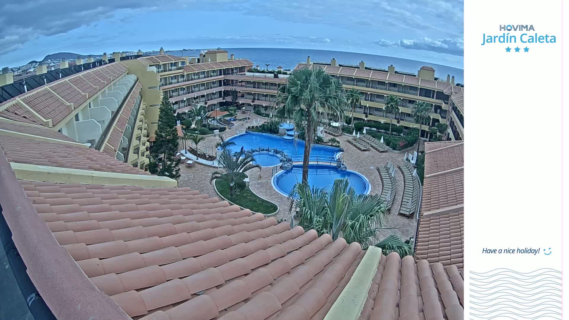 A tan-tiled roof overlooks a courtyard with a swimming pool and apartment buildings, under a partly cloudy sky.