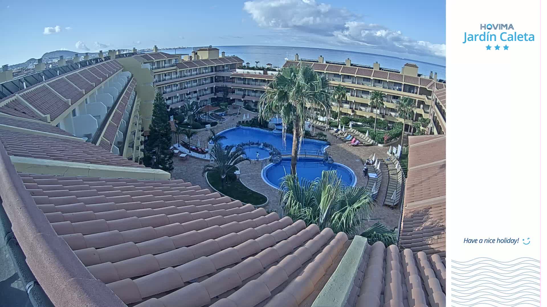 A resort with a courtyard featuring two swimming pools is viewed from an adjacent building's roof under a partly cloudy sky.