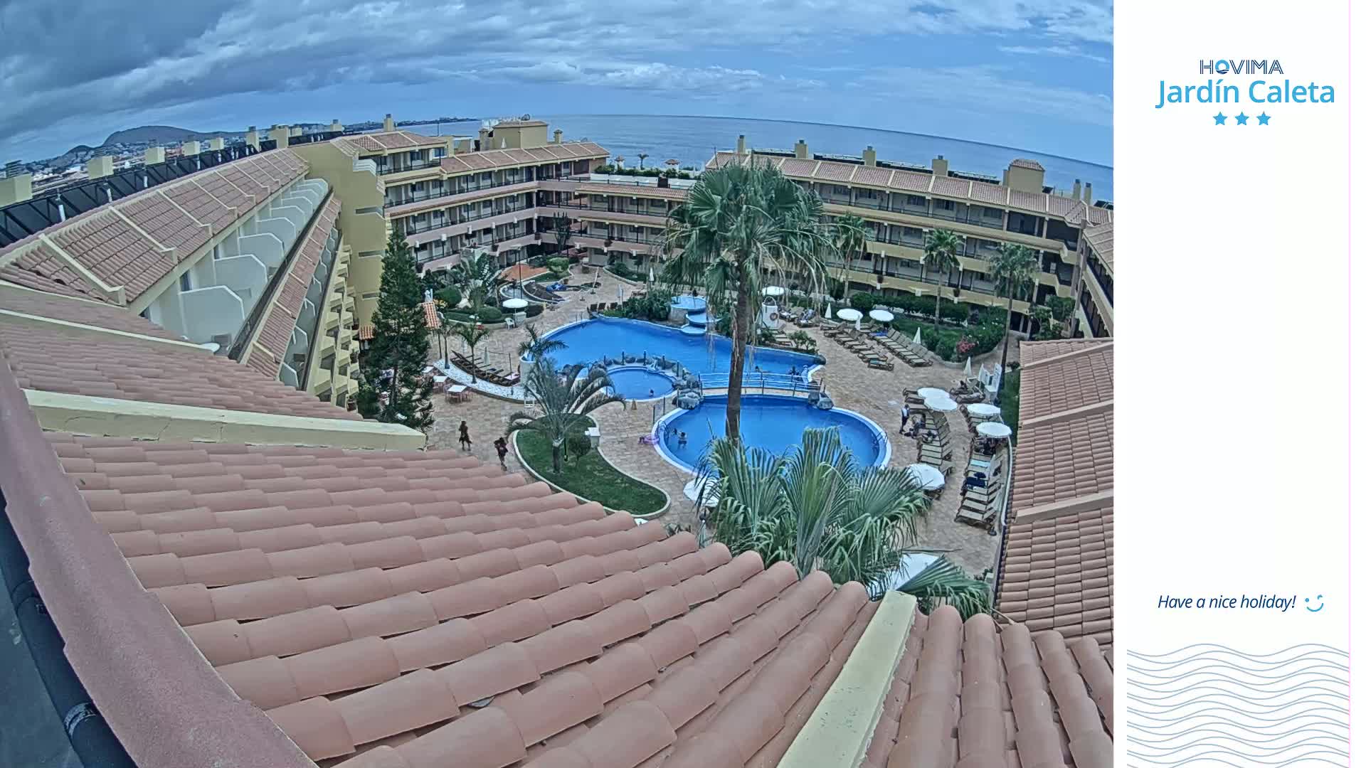 An overhead view shows a resort with multiple buildings surrounding a pool area on a partly cloudy day.