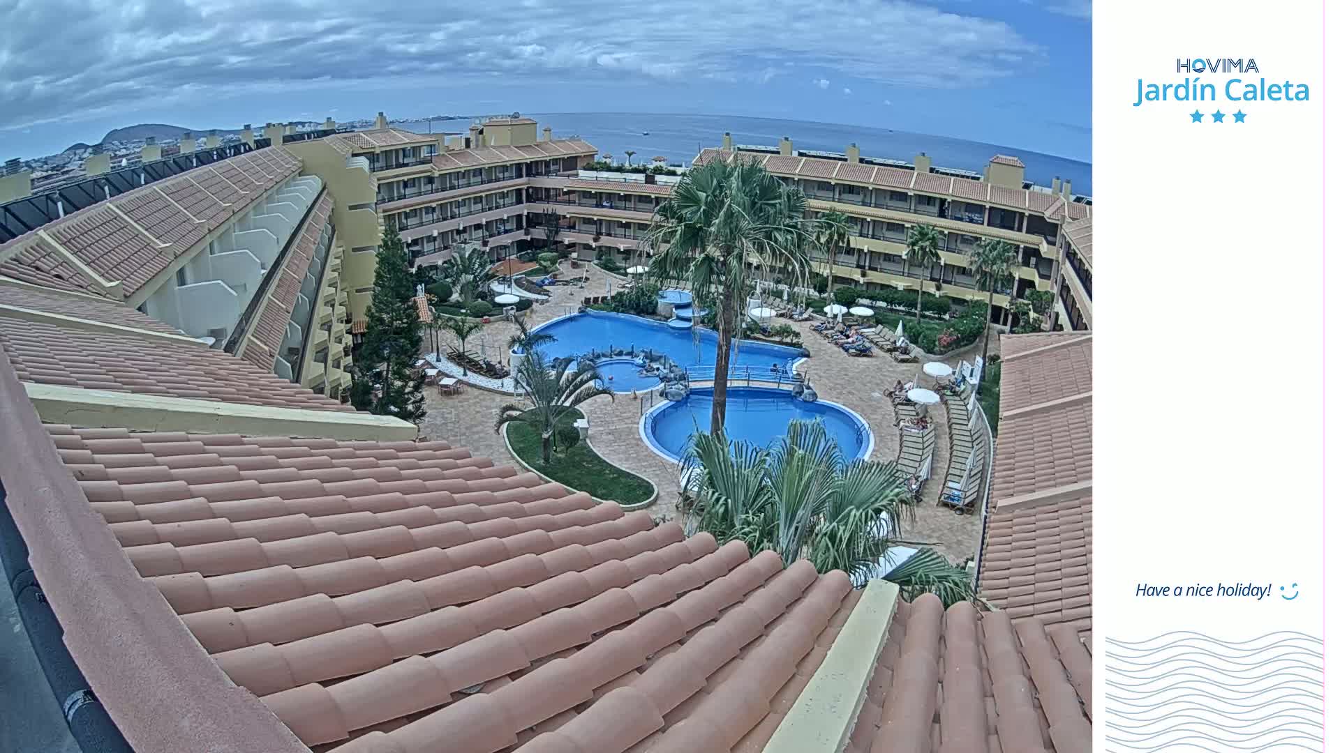 An aerial view shows a resort with two swimming pools surrounded by buildings and palm trees under a cloudy sky.