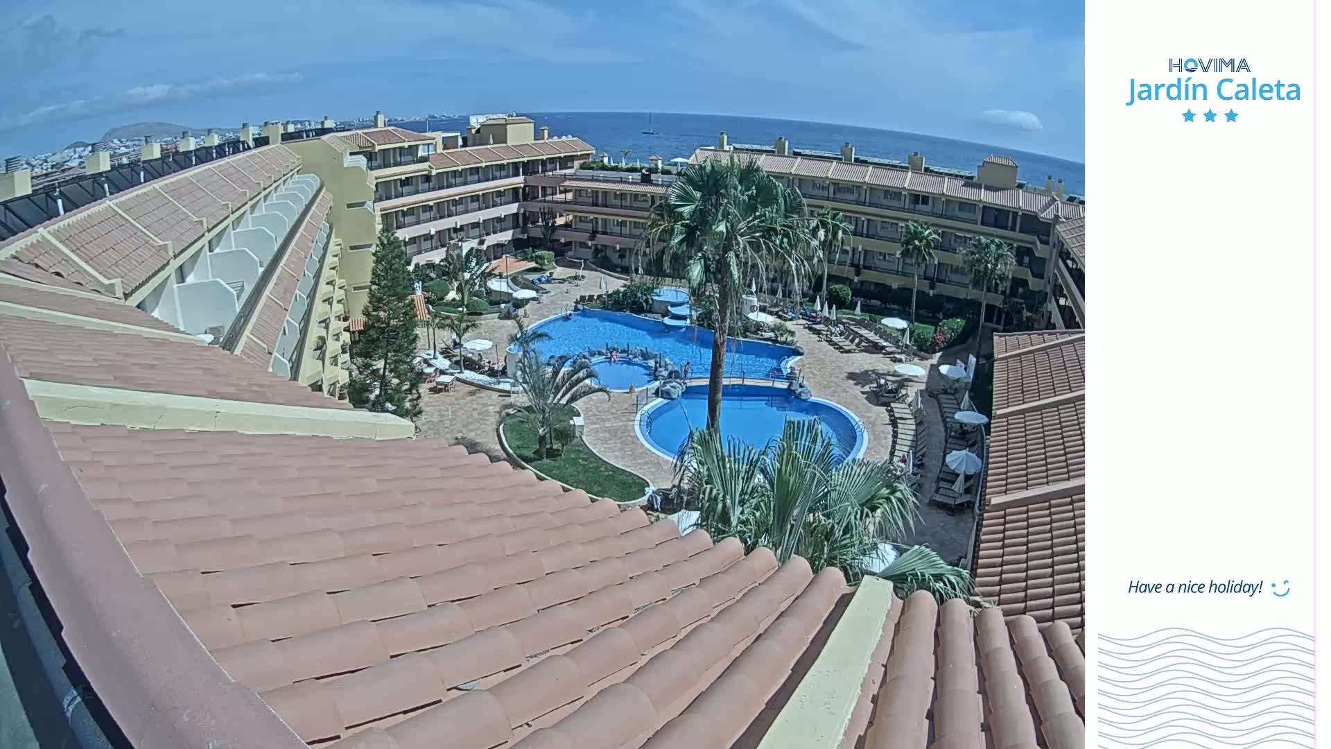A high-angle view shows a resort complex with multiple pools surrounded by buildings and palm trees under sunny skies and a clear ocean in the background.