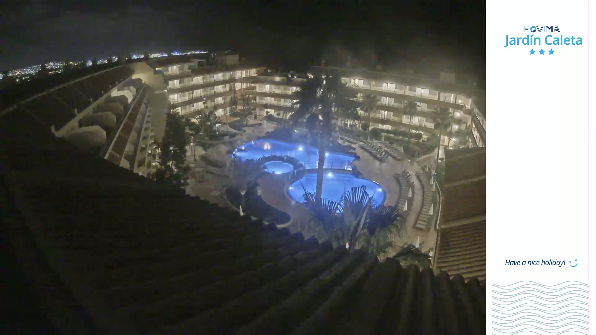 A nighttime aerial view shows a multi-story building surrounding a complex of illuminated swimming pools.