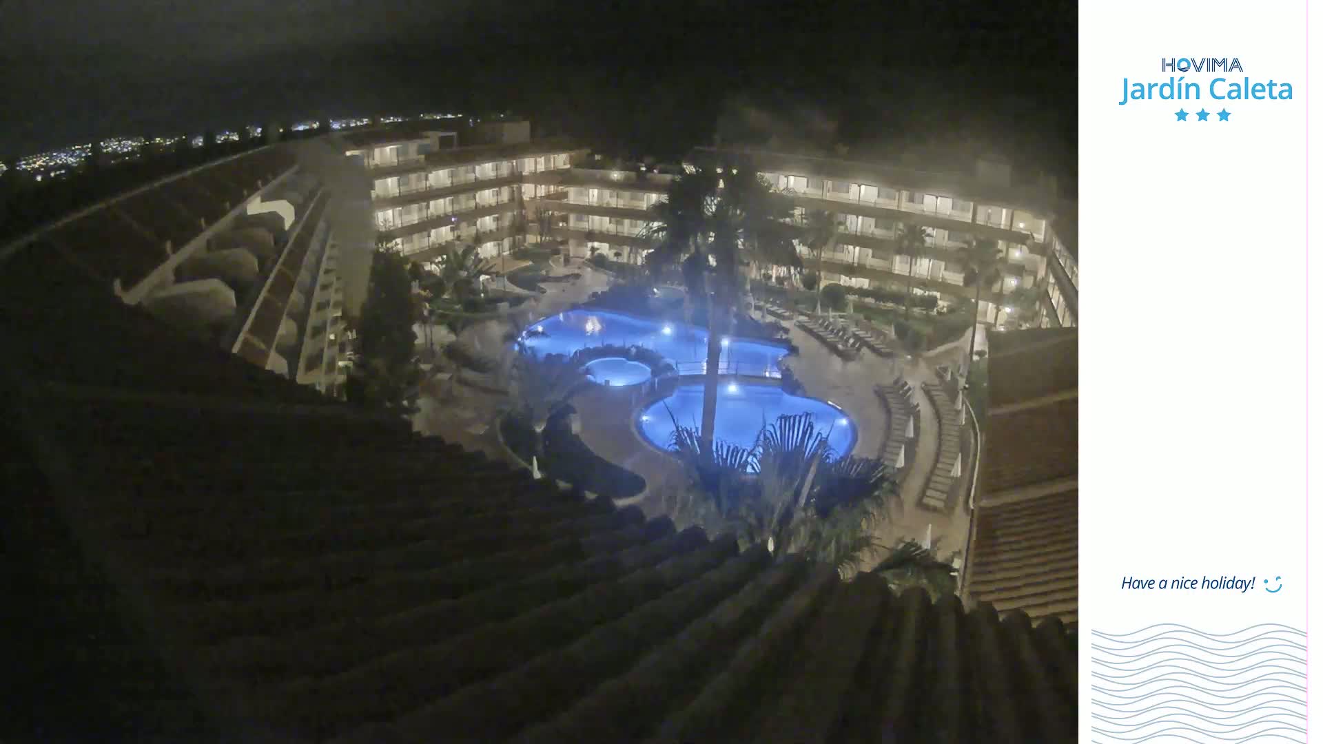 A nighttime, high-angle view shows a multi-story building complex surrounding a lit swimming pool area.