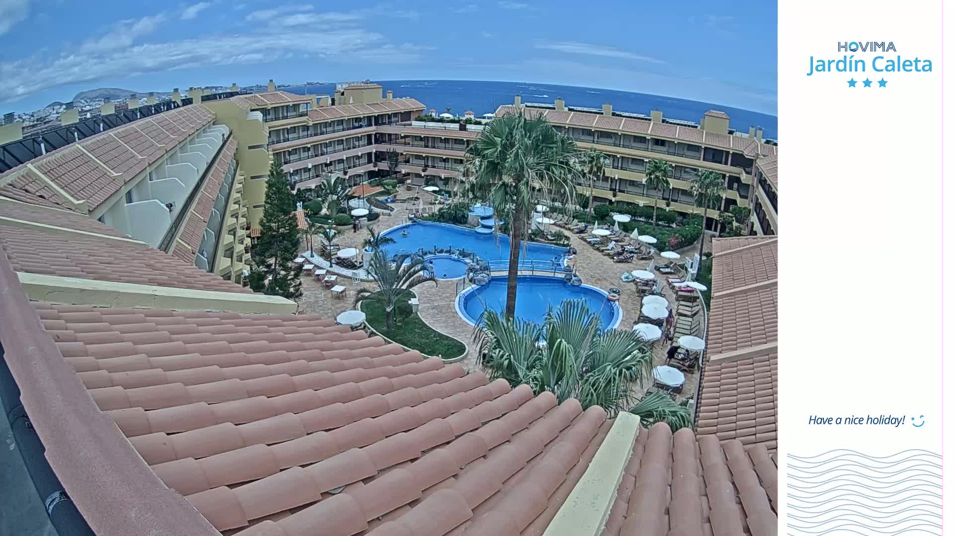 A partly sunny aerial view shows a resort complex with multiple buildings surrounding a swimming pool area, and an ocean view in the background.