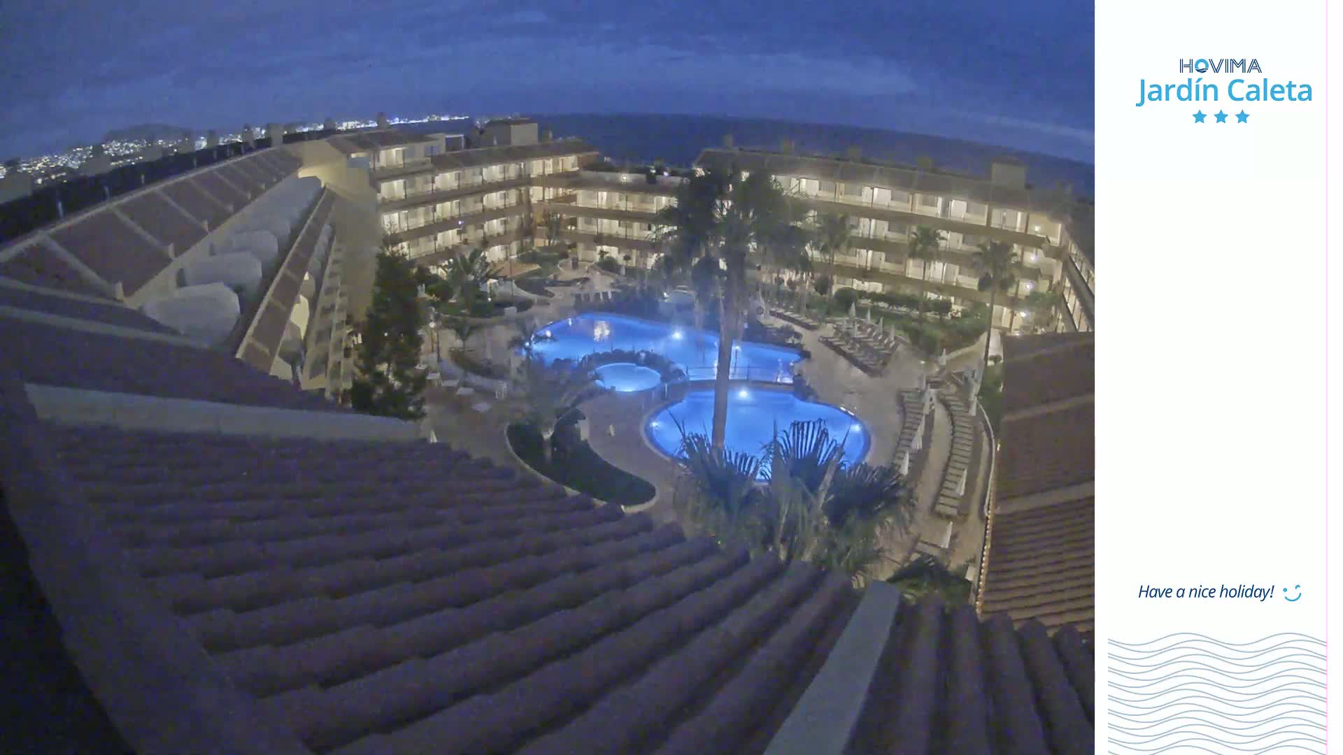 A nighttime aerial view shows a multi-story resort building surrounding illuminated swimming pools.