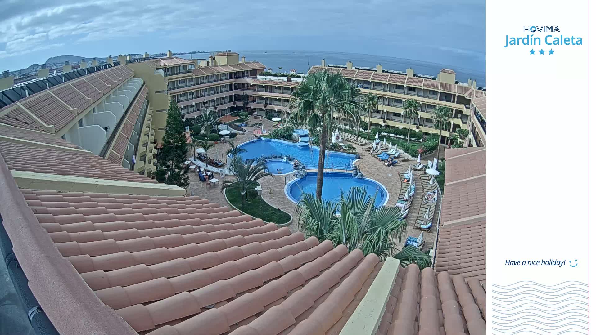 An overhead view of a resort with two swimming pools surrounded by buildings under a partly cloudy sky.