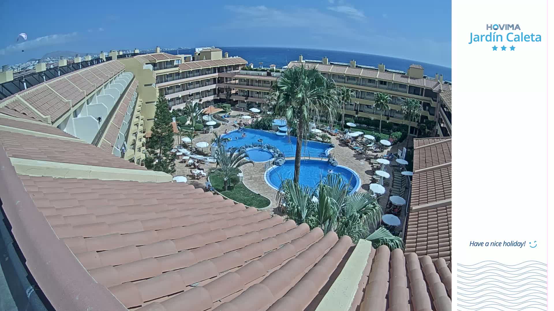 A tiled roof overlooks a sunny resort pool area with several palm trees and lounge chairs, all situated near a clear blue ocean under a bright, cloudless sky.