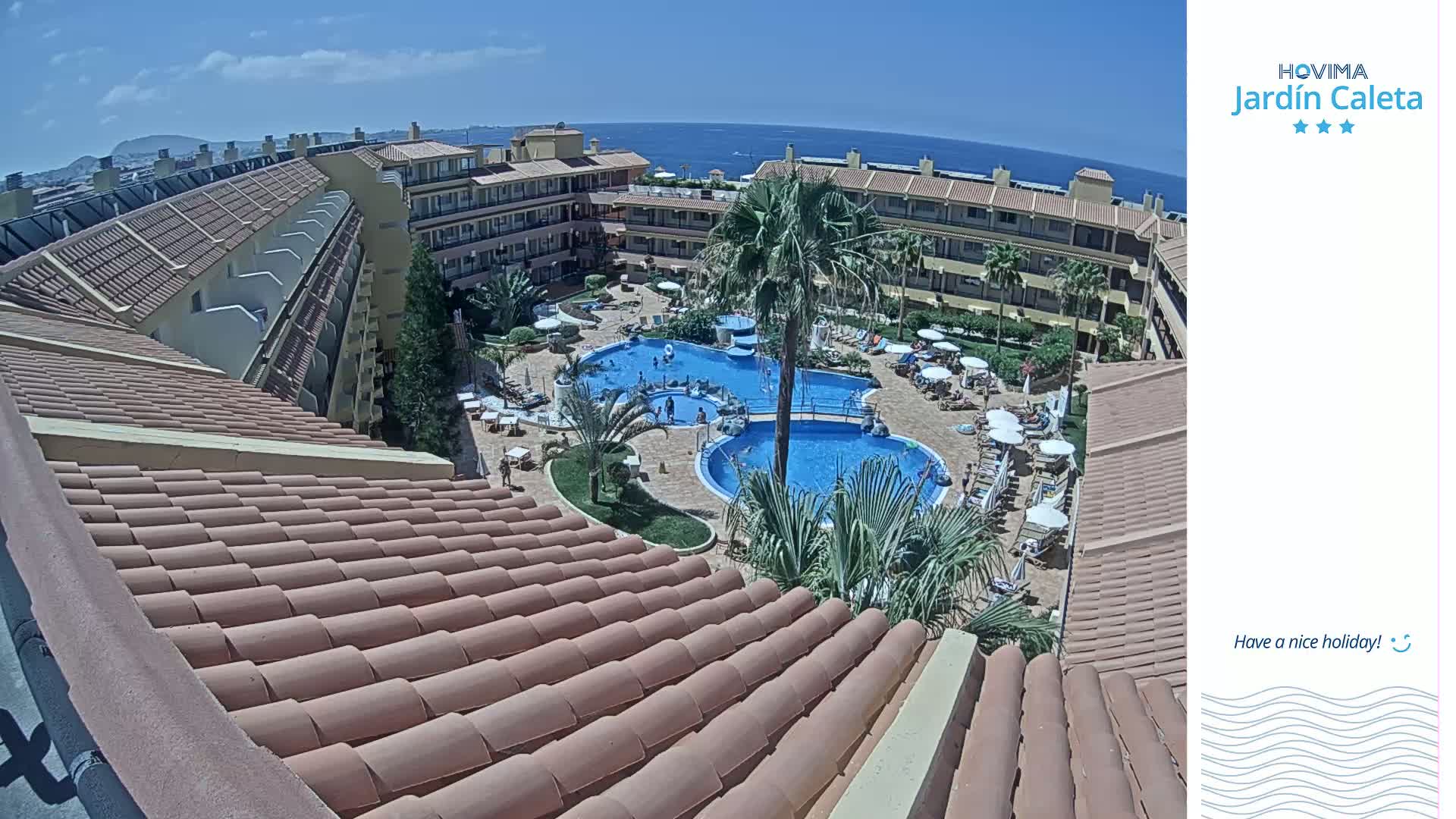 A rooftop overlooks a sunny courtyard with a swimming pool and surrounding buildings, the ocean visible in the distance.