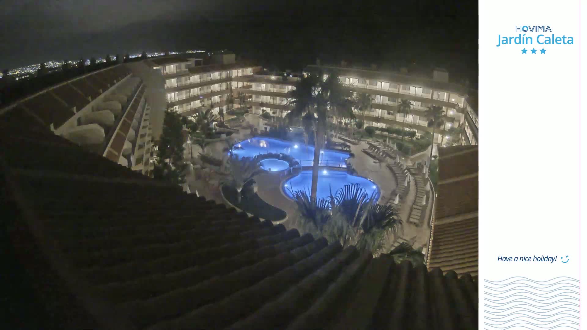 A nighttime, high-angle view shows a multi-story building complex surrounding illuminated swimming pools, with palm trees and lounge chairs visible in the courtyard.