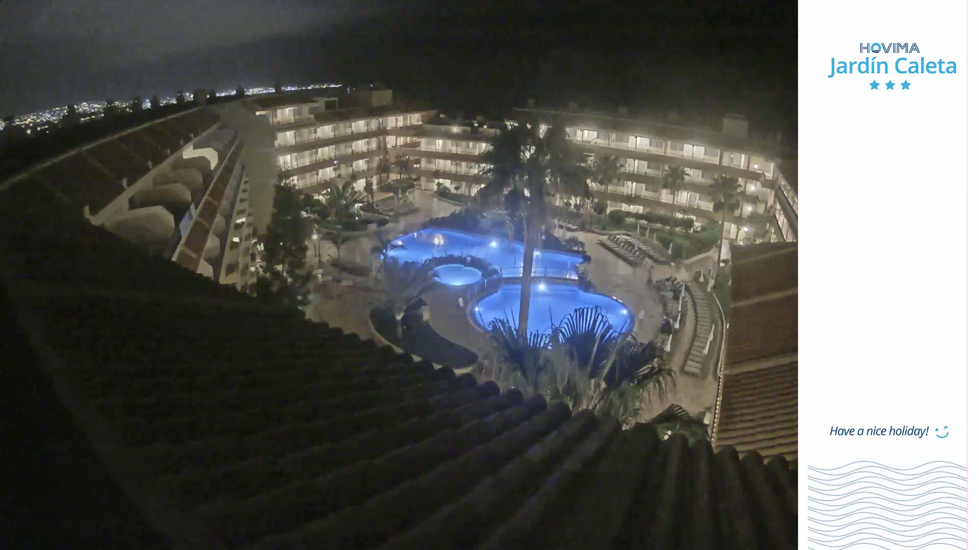 A nighttime view from a rooftop shows a multi-story building surrounding a complex of illuminated pools and landscaping.