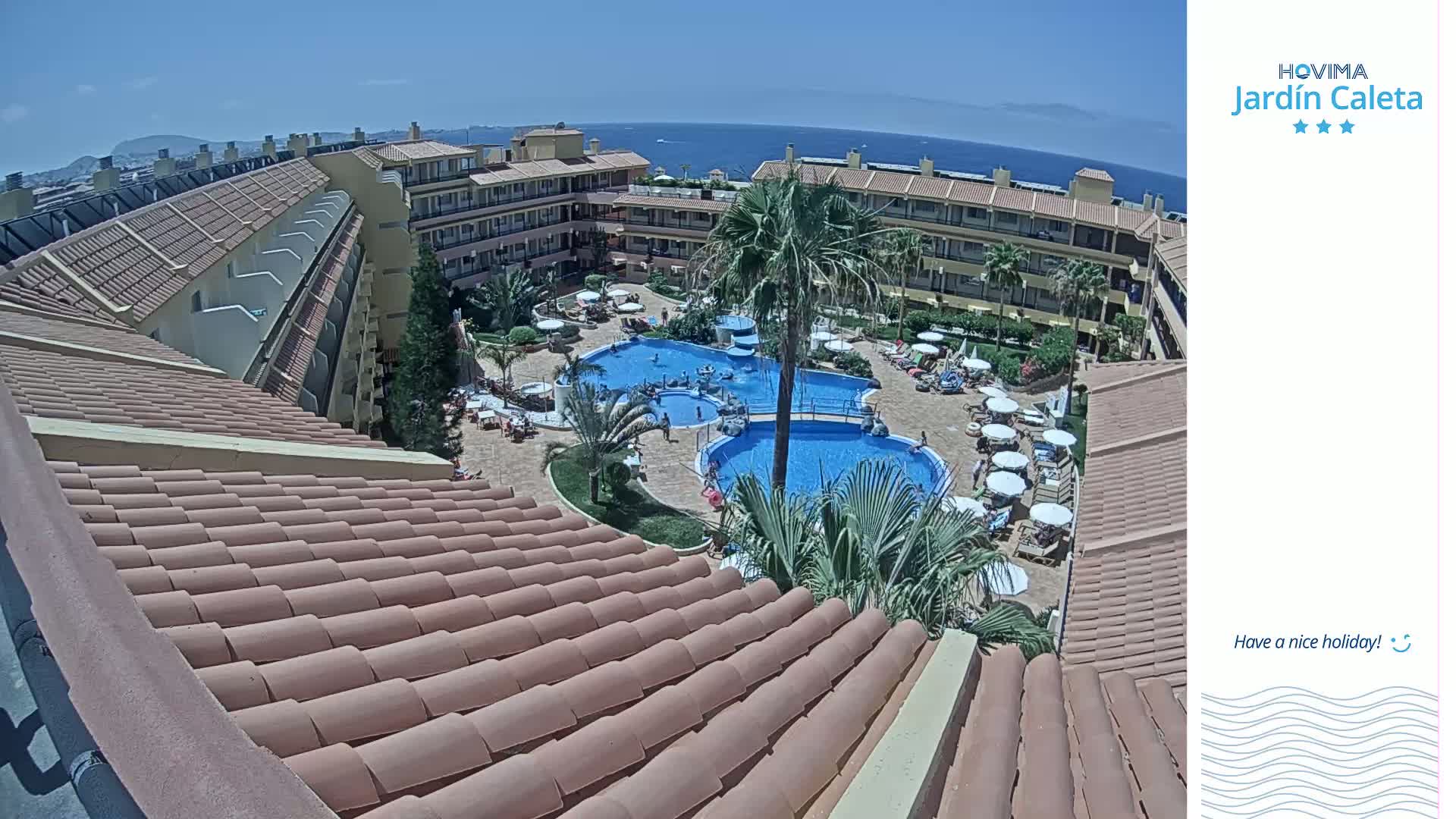 An overhead view of a resort complex with multiple swimming pools, surrounded by buildings and palm trees, on a sunny day with a clear view of the ocean in the distance.