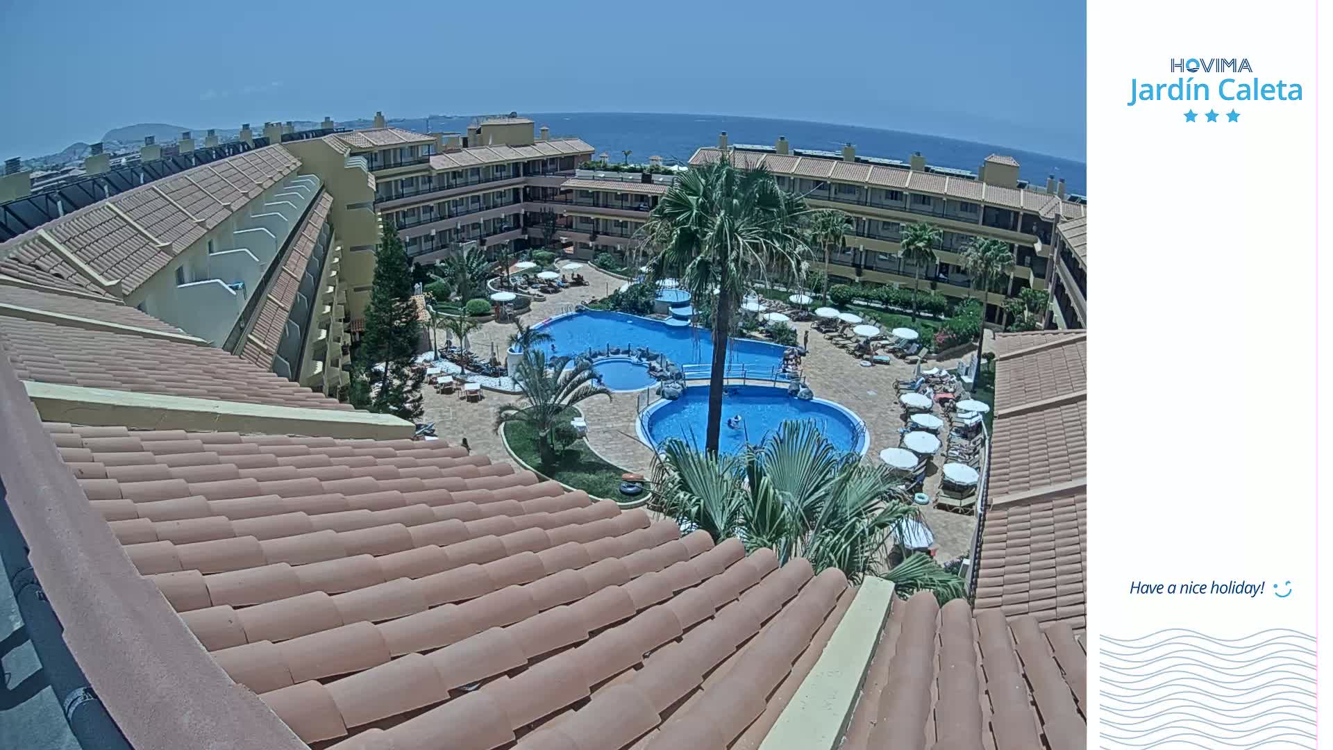 An overhead view of a resort complex with multiple swimming pools and surrounding buildings on a sunny day, overlooking the ocean.