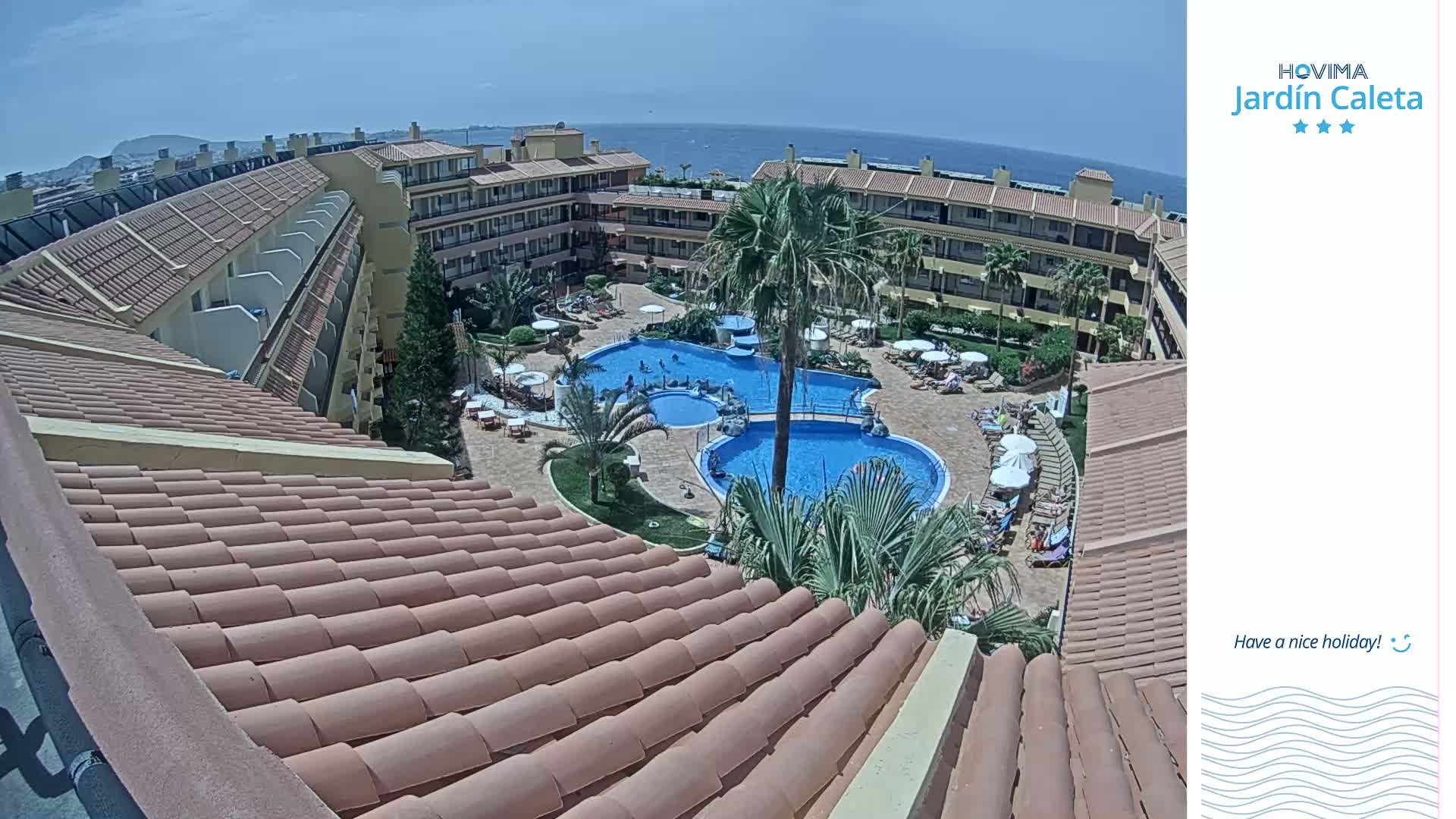 An aerial view shows a resort complex with multiple buildings surrounding two swimming pools on a sunny day with a clear blue ocean in the distance.