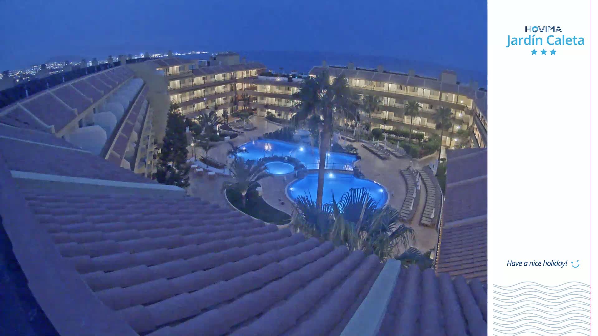 An aerial nighttime view shows a resort with multiple pools illuminated in blue, surrounded by low-rise buildings under a dark blue sky.
