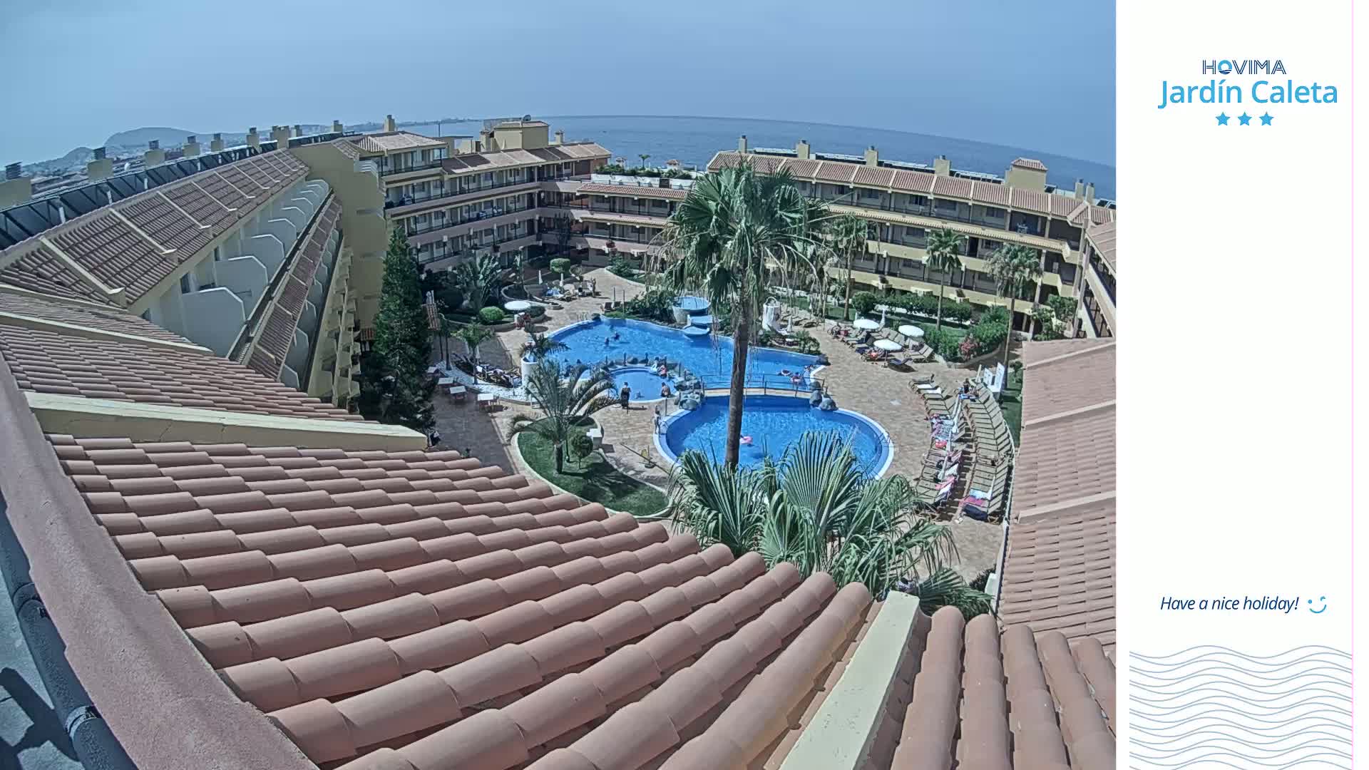 An aerial view shows a resort complex with multiple buildings surrounding a large swimming pool and a smaller children's pool, on a sunny day with a clear blue ocean in the distance.