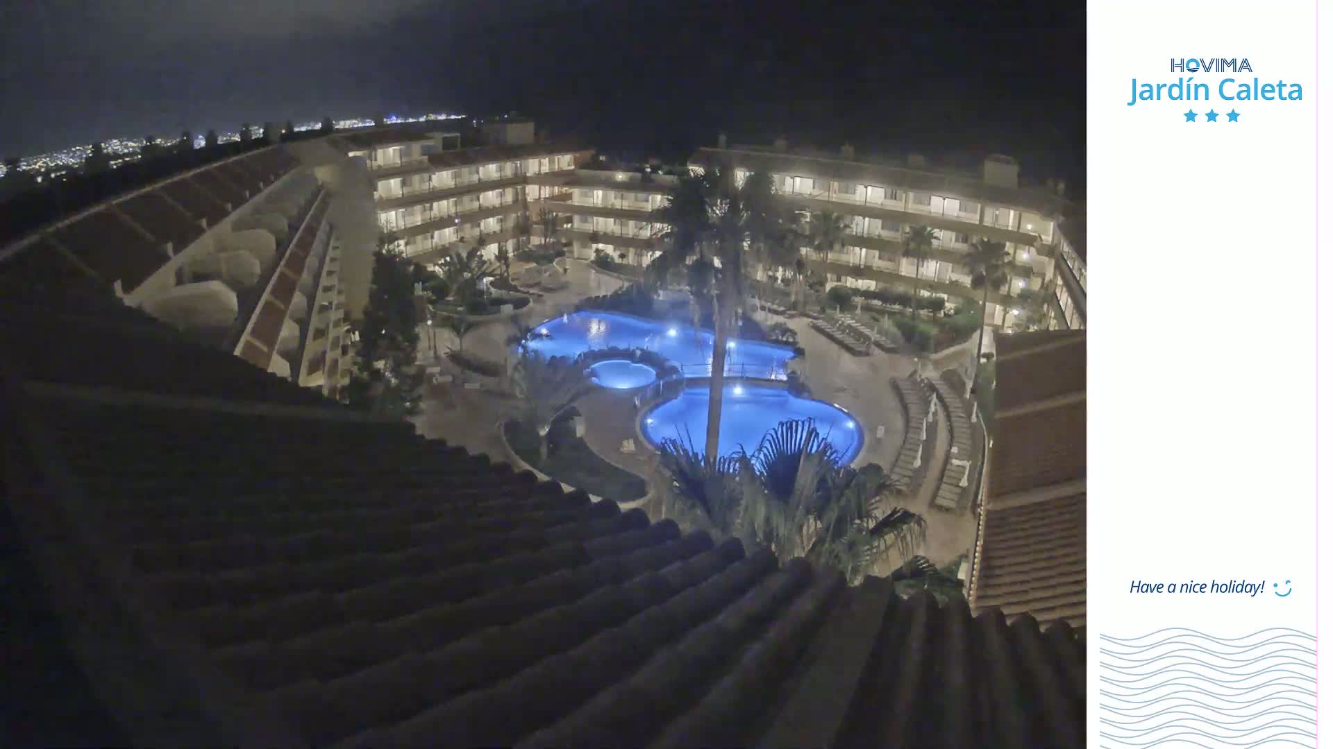 A nighttime, overhead view shows a multi-story building complex surrounding a lit swimming pool area with palm trees.