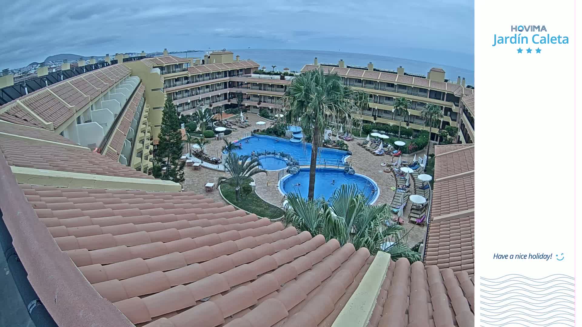 An overhead view of a resort with multiple pools, surrounded by buildings and palm trees, under an overcast sky.