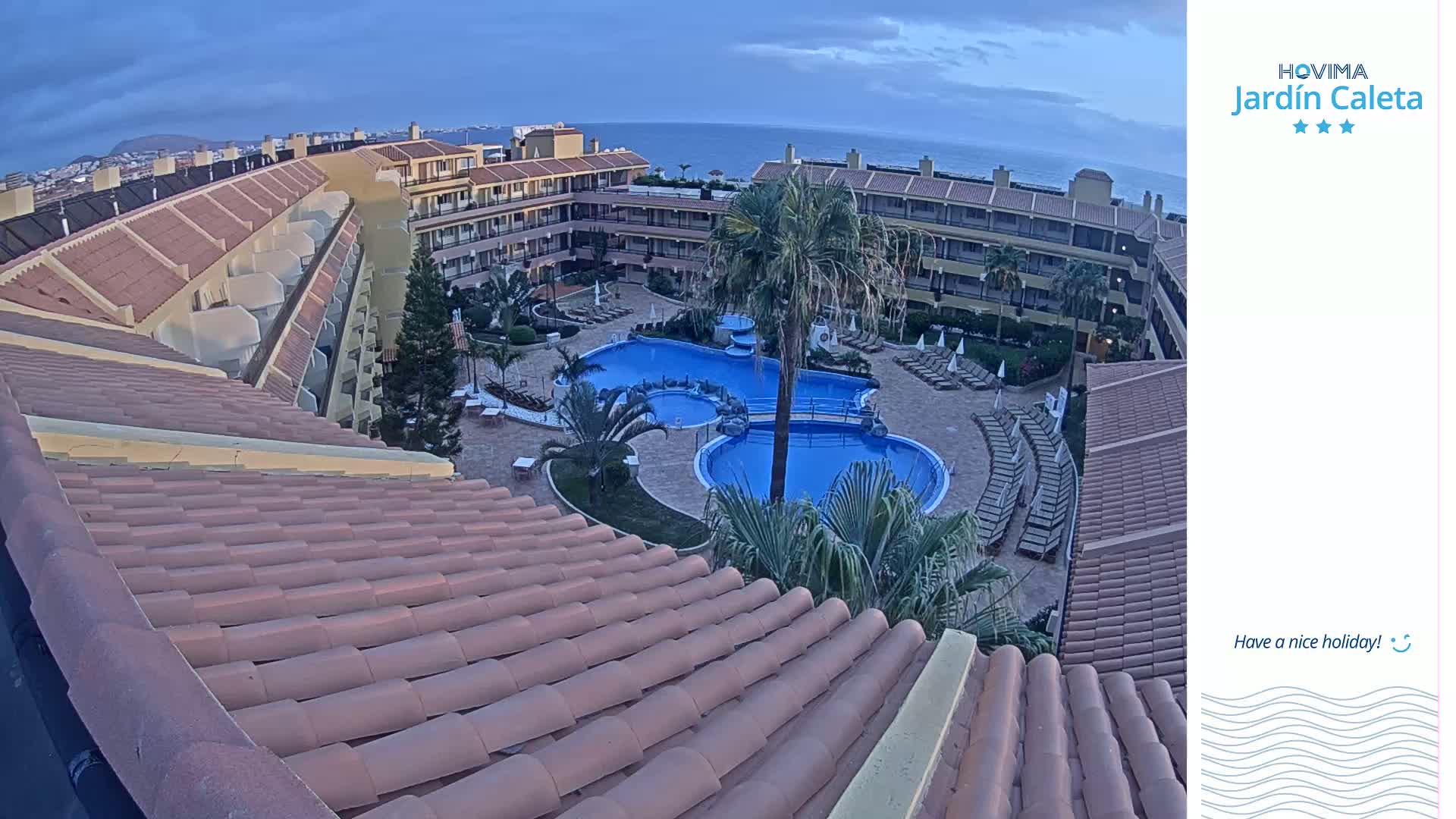An aerial view shows a resort with a large swimming pool surrounded by buildings under a partly cloudy sky.