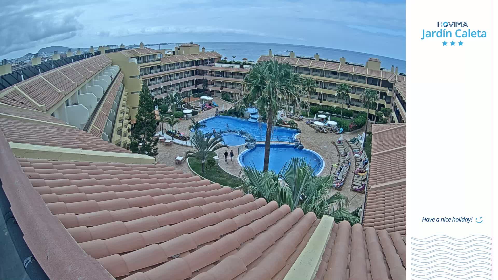 An overhead view of a resort pool area with multiple buildings and a tiled roof in the foreground, under an overcast sky.
