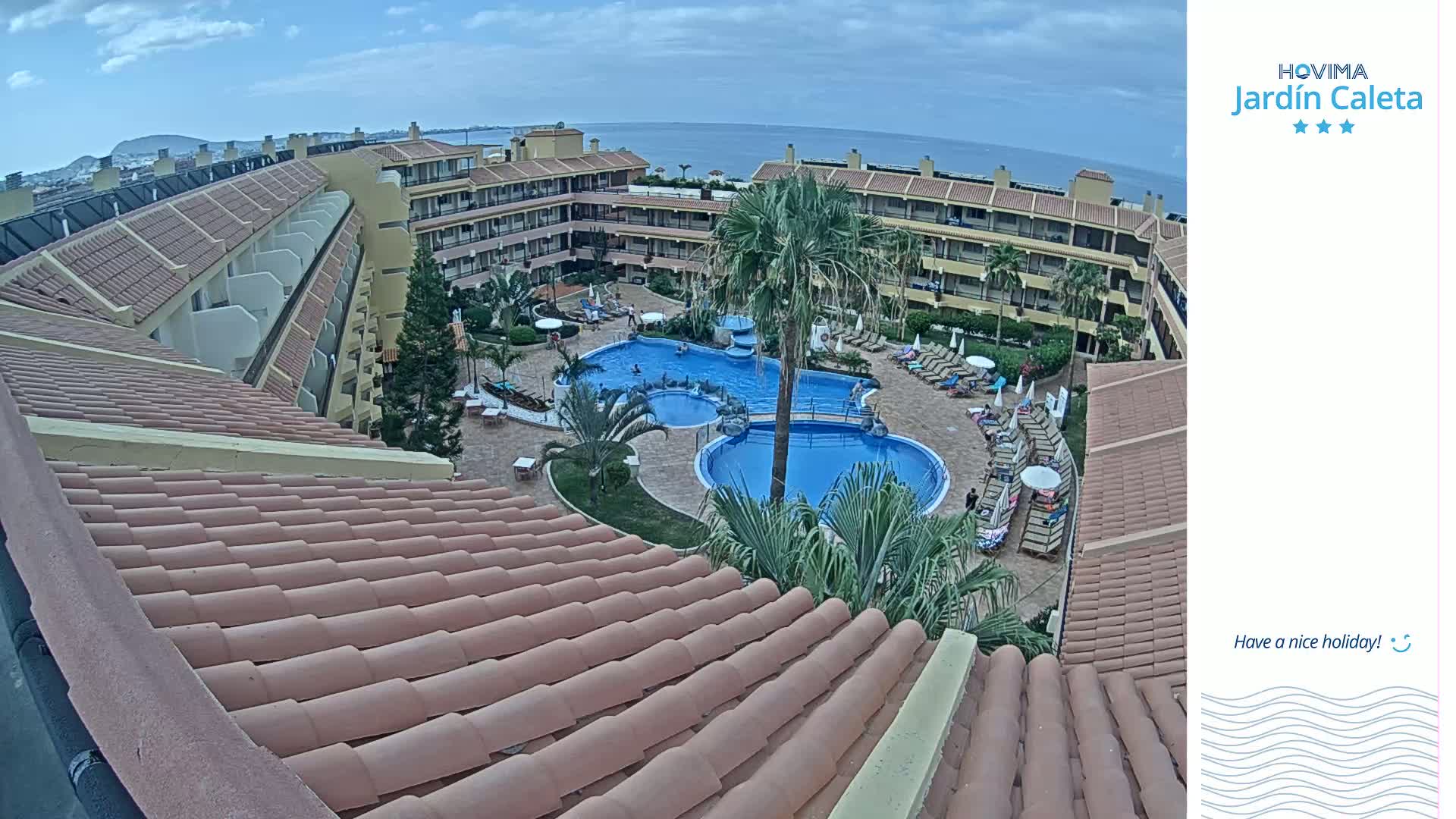 An aerial view shows a resort pool area with several people swimming and lounging on chairs under a partly cloudy sky.