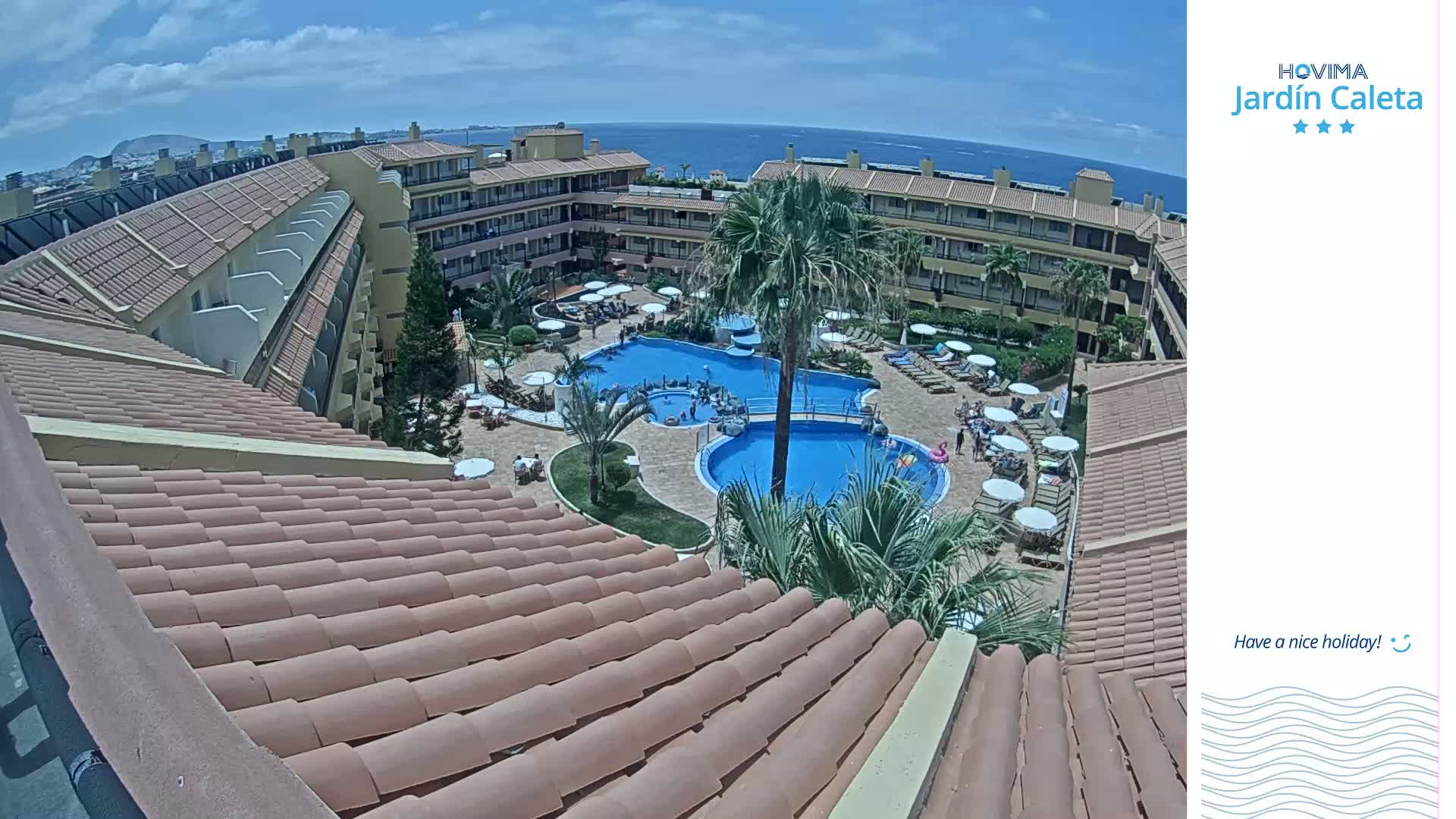 An overhead view of a resort's pool area on a sunny day shows multiple pools surrounded by lounge chairs and palm trees, with a building's tiled roof in the foreground.
