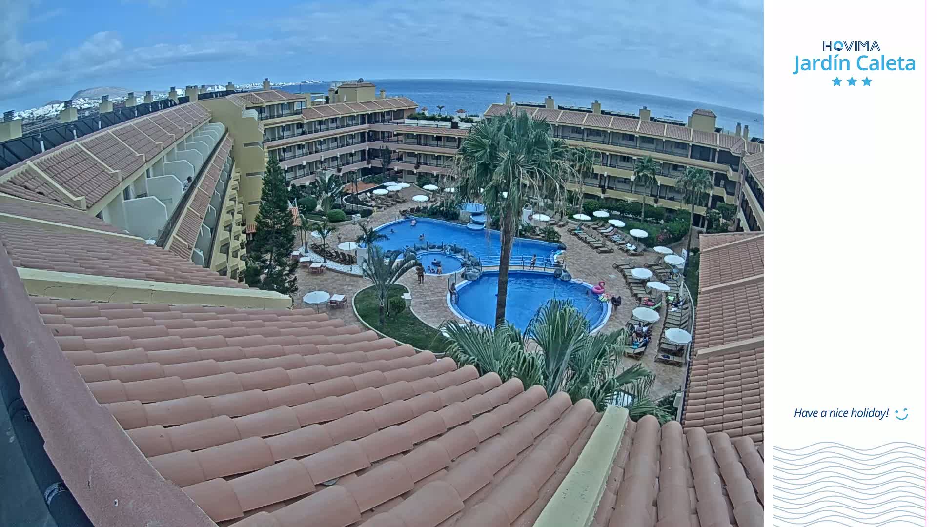 An overhead view shows a resort complex with a large pool and surrounding buildings under a partly cloudy sky.