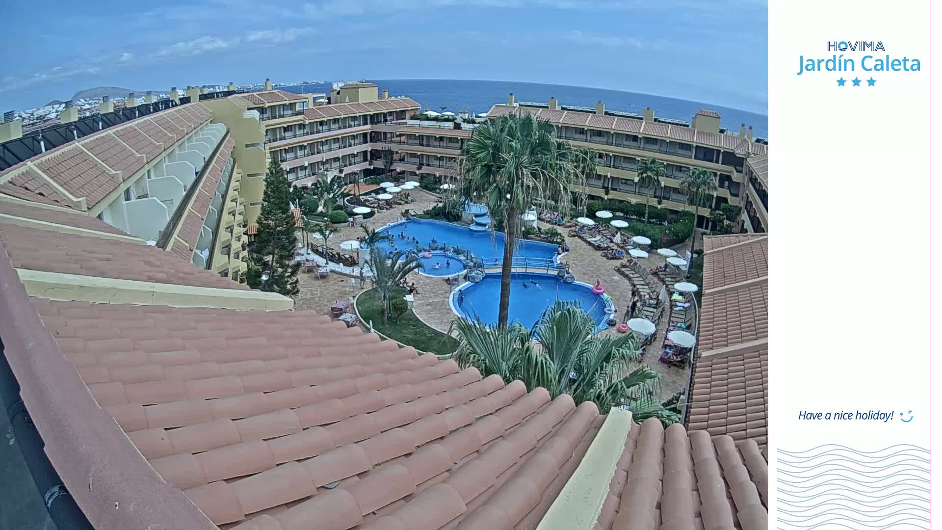 An overhead view shows a multi-story building with a tiled roof surrounding a courtyard with two swimming pools, palm trees, and many people enjoying a sunny day.