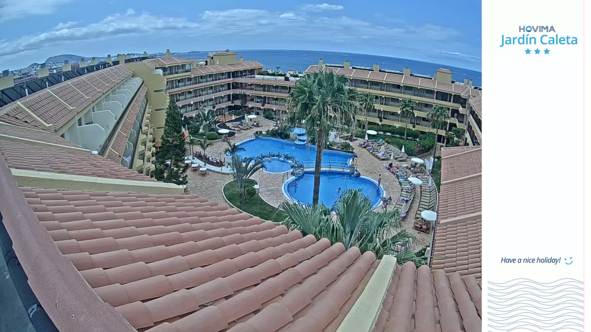 An overhead view shows a resort pool area with several people swimming and lounging on chairs, under a partly cloudy sky and overlooking a calm ocean.