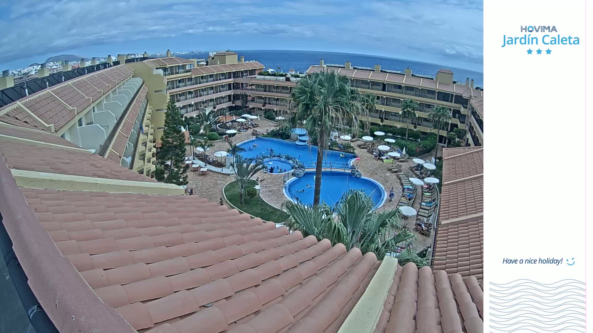 An overhead view of a resort pool area with multiple pools, surrounded by buildings under a partly cloudy sky.