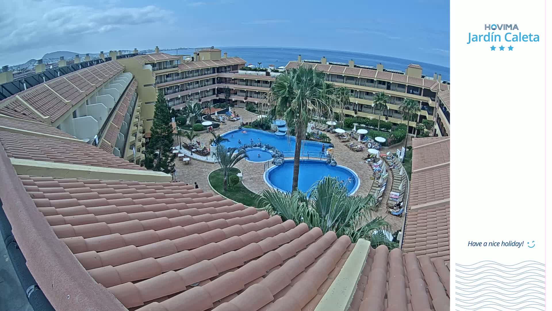 An aerial view of a resort pool area with multiple buildings and a tiled roof in the foreground, under a partly cloudy sky.
