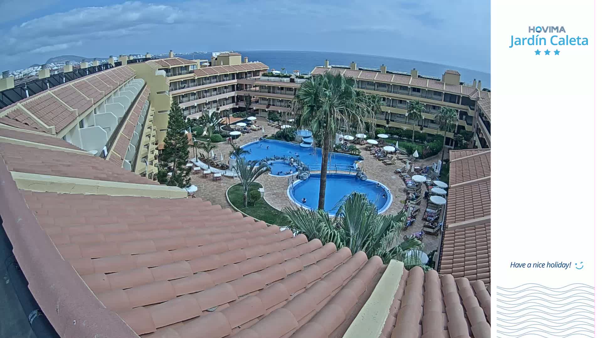 An elevated view of a resort pool area with multiple pools surrounded by buildings under a partly cloudy sky.
