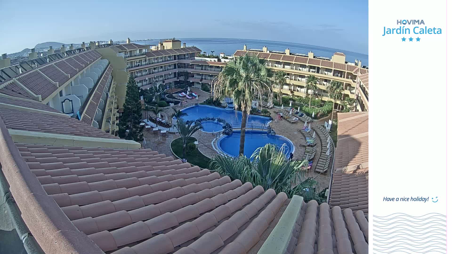 An aerial view shows a resort with a multi-level building surrounding a swimming pool complex on a sunny day, overlooking a body of water in the distance.