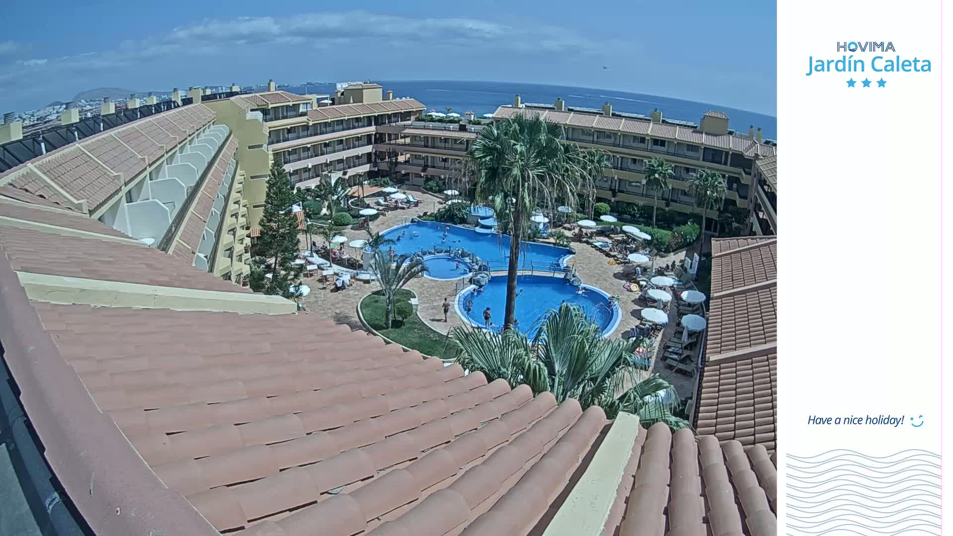An overhead view of a resort pool area with multiple pools surrounded by palm trees and buildings under a sunny sky, overlooking the ocean in the distance.