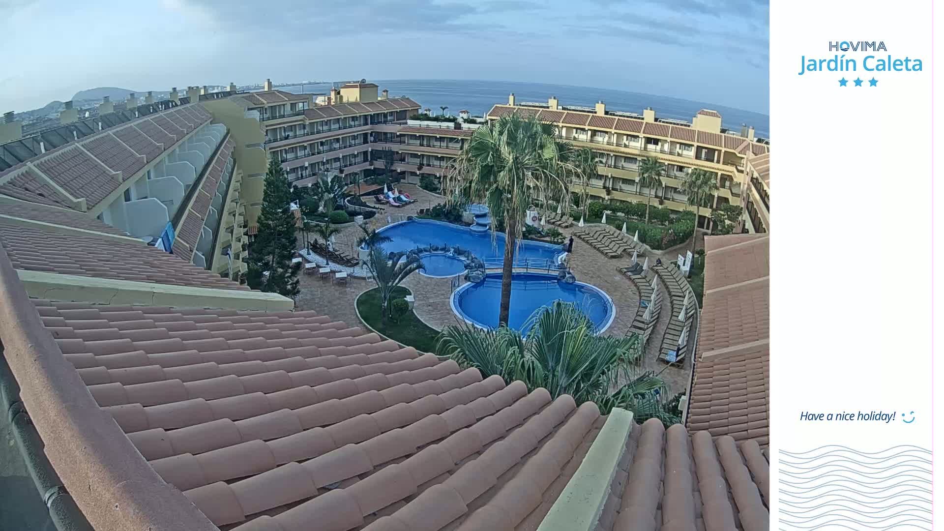 An overhead view of a resort complex with a large pool, surrounded by buildings under a partly cloudy sky.