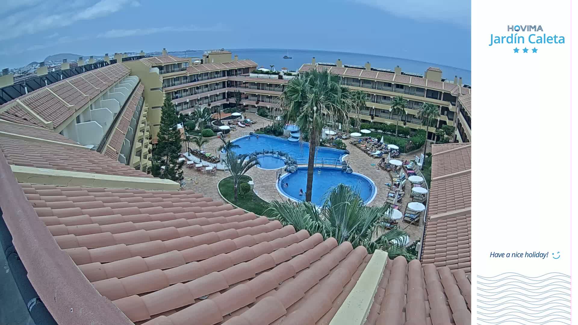 A resort with a large pool and surrounding buildings is viewed from an elevated position on a partly cloudy day.