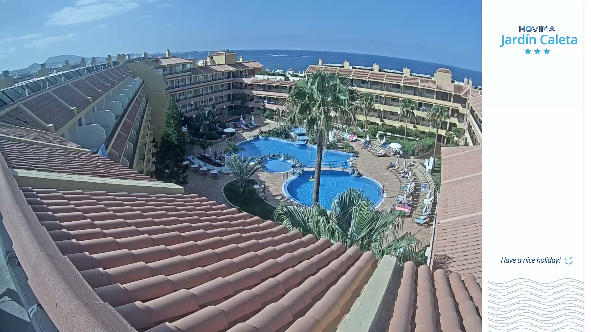 An aerial view of a resort courtyard with two swimming pools, sunbathing areas, and surrounding buildings on a sunny day.