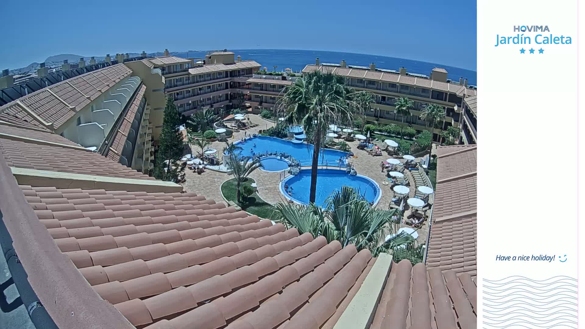 A tiled roof overlooks a sunny courtyard with a swimming pool and surrounding buildings near the ocean.